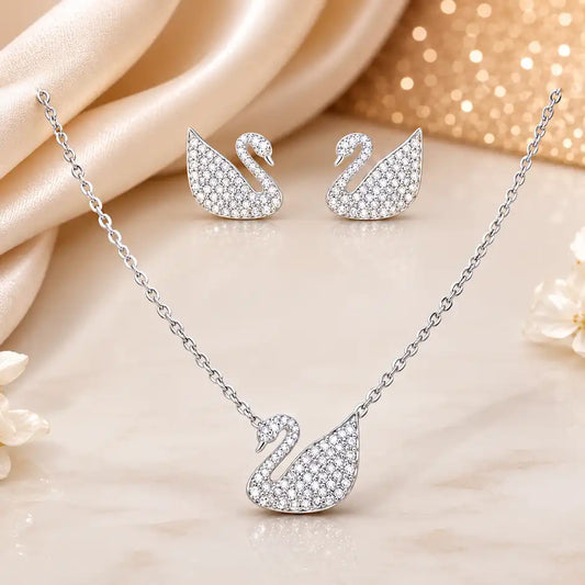 Silver-toned swan necklace and matching stud earrings set with pavé clear stones, arranged on a soft beige marble surface with draped satin fabric and a warm gold glitter backdrop.