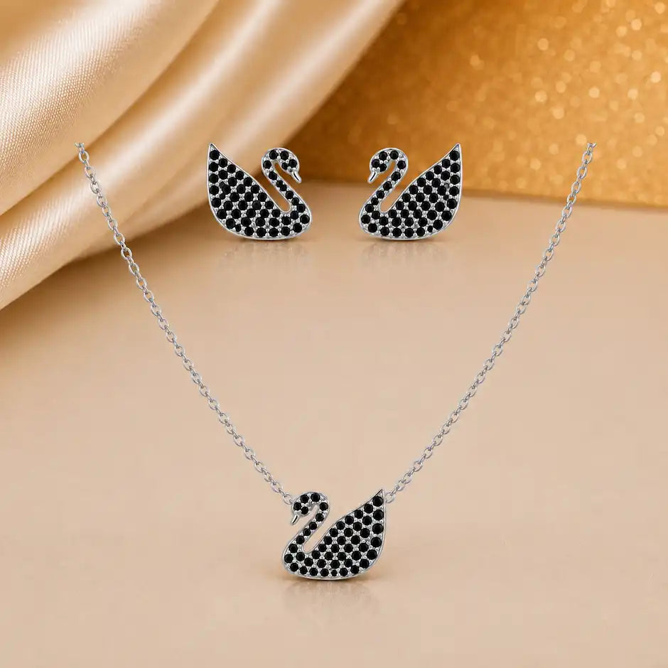 A three-piece jewelry set featuring a silver-toned necklace, matching stud earrings, and a swan-shaped pendant, all embellished with small black stones, displayed on a beige surface with draped satin fabric.