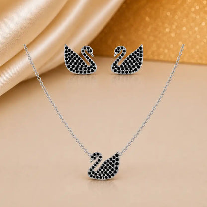 A three-piece jewelry set featuring a silver-toned necklace, matching stud earrings, and a swan-shaped pendant, all embellished with small black stones, displayed on a beige surface with draped satin fabric.