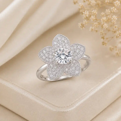 Silver or white gold flower-shaped ring featuring a vibrant, round-cut blue sapphire center stone surrounded by five pavé-set diamond petals, displayed on a soft satin background.