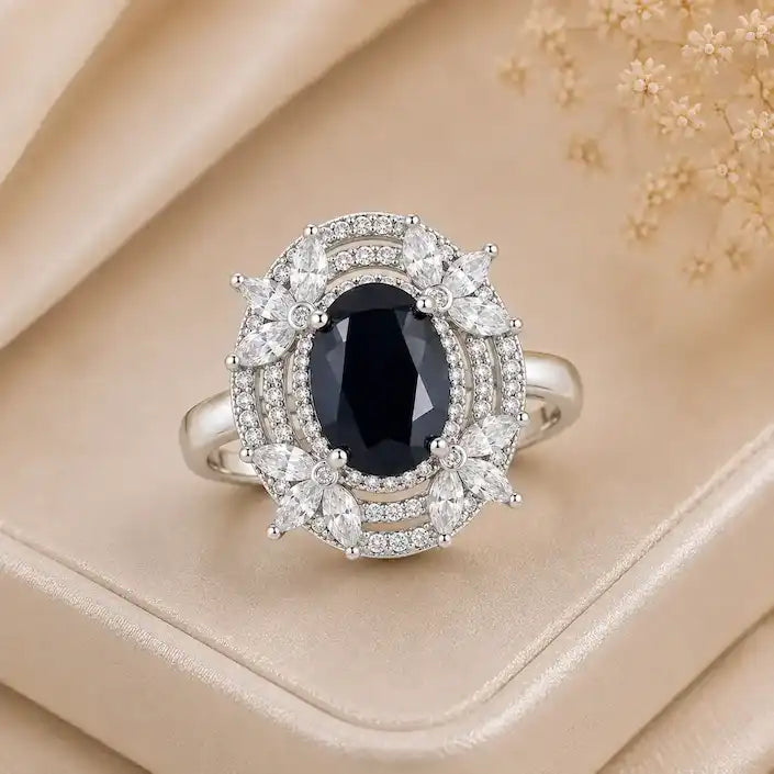 Elegant silver ring featuring a deep blue oval gemstone, surrounded by sparkling halo details and delicate marquise-shaped crystal accents, displayed on a cream satin jewelry box.