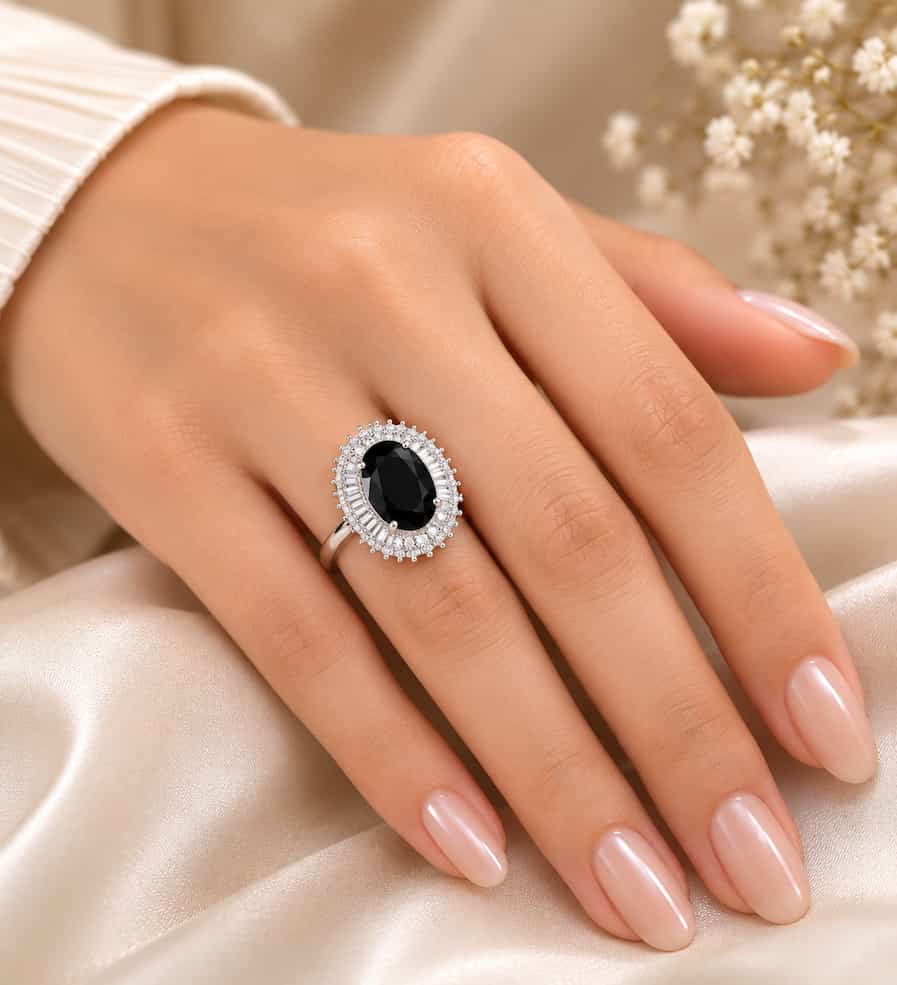 Elegant silver ring featuring a large oval black gemstone surrounded by a sparkling halo of clear stones, displayed on a cream satin jewelry box with delicate white flowers in the background.