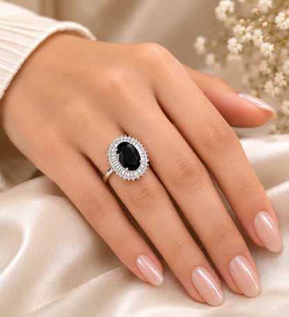 Elegant silver ring featuring a large oval black gemstone surrounded by a sparkling halo of clear stones, displayed on a cream satin jewelry box with delicate white flowers in the background.