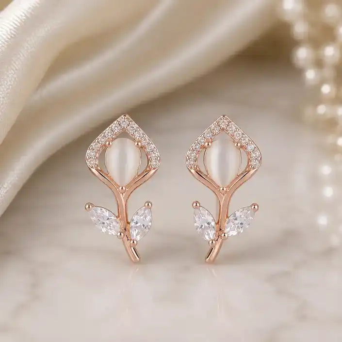 Rose gold stud earrings shaped like delicate flower buds, featuring an oval cat's eye stone centerpiece framed by pavé crystals, with marquise-cut crystal leaves on the stem, displayed on a marble surface.