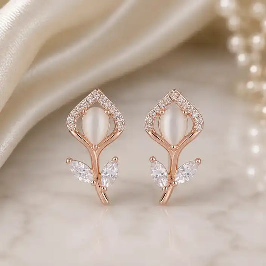 Rose gold stud earrings shaped like delicate flower buds, featuring an oval cat's eye stone centerpiece framed by pavé crystals, with marquise-cut crystal leaves on the stem, displayed on a marble surface.