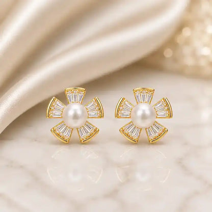 A pair of gold flower stud earrings featuring a round white pearl center surrounded by five fan-shaped petals, each set with tapered baguette crystals and small pavé crystal accents, displayed on a reflective marble surface against a soft champagne satin background.