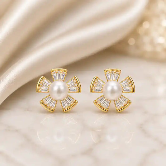 A pair of gold flower stud earrings featuring a round white pearl center surrounded by five fan-shaped petals, each set with tapered baguette crystals and small pavé crystal accents, displayed on a reflective marble surface against a soft champagne satin background.