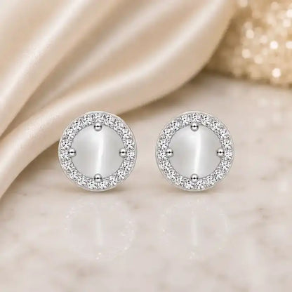 A pair of round gold stud earrings featuring a circular, milky white cat's eye stone centerpiece held by four small prongs, encircled by a halo of shimmering pavé crystals, displayed on a polished white marble surface with a draped champagne satin background.
