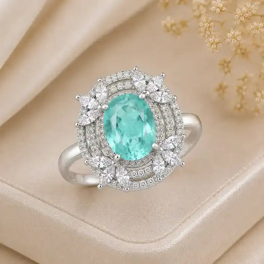 Elegant silver ring featuring a deep blue oval gemstone, surrounded by sparkling halo details and delicate marquise-shaped crystal accents, displayed on a cream satin jewelry box.